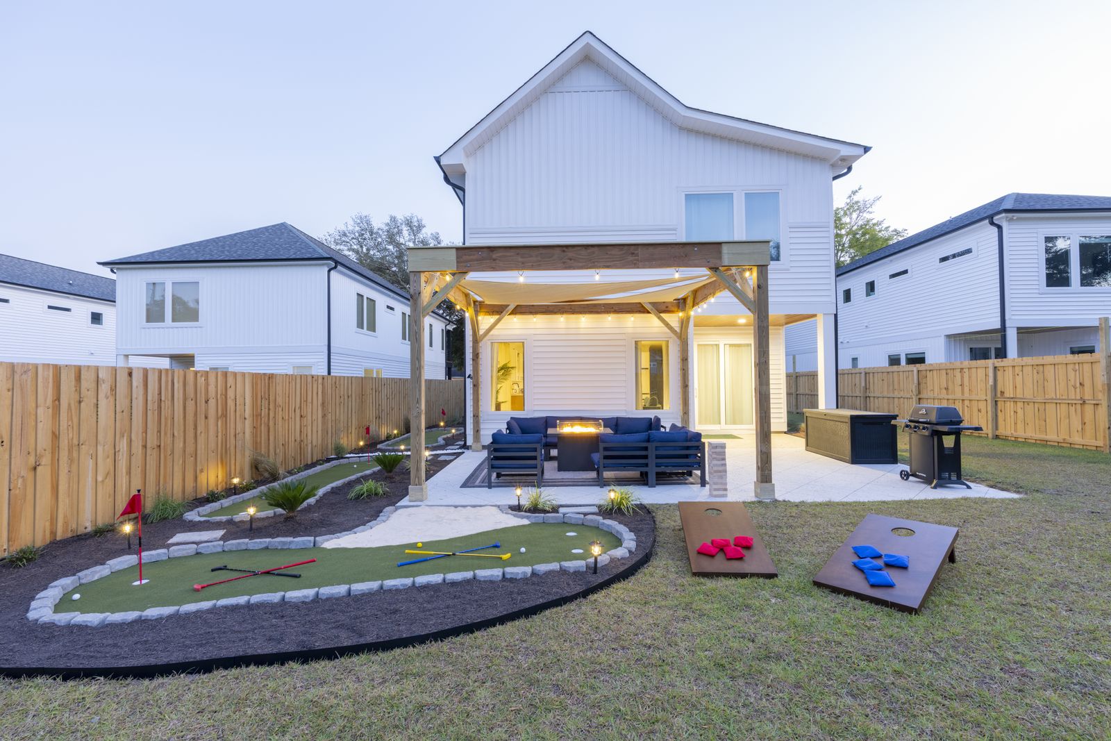 The backyard at dusk with a lit pergola over an outdoor lounge and fire table, with putting greens and cornhole on the lawn