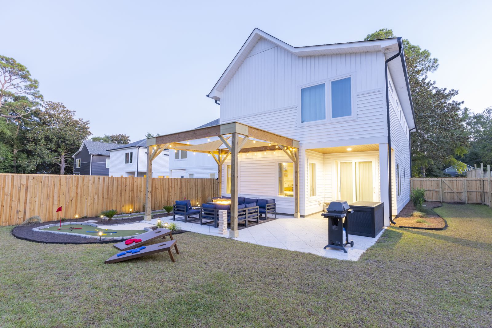 Twilight view of the back of the house with a lit pergola, outdoor sectional, grill, and cornhole boards