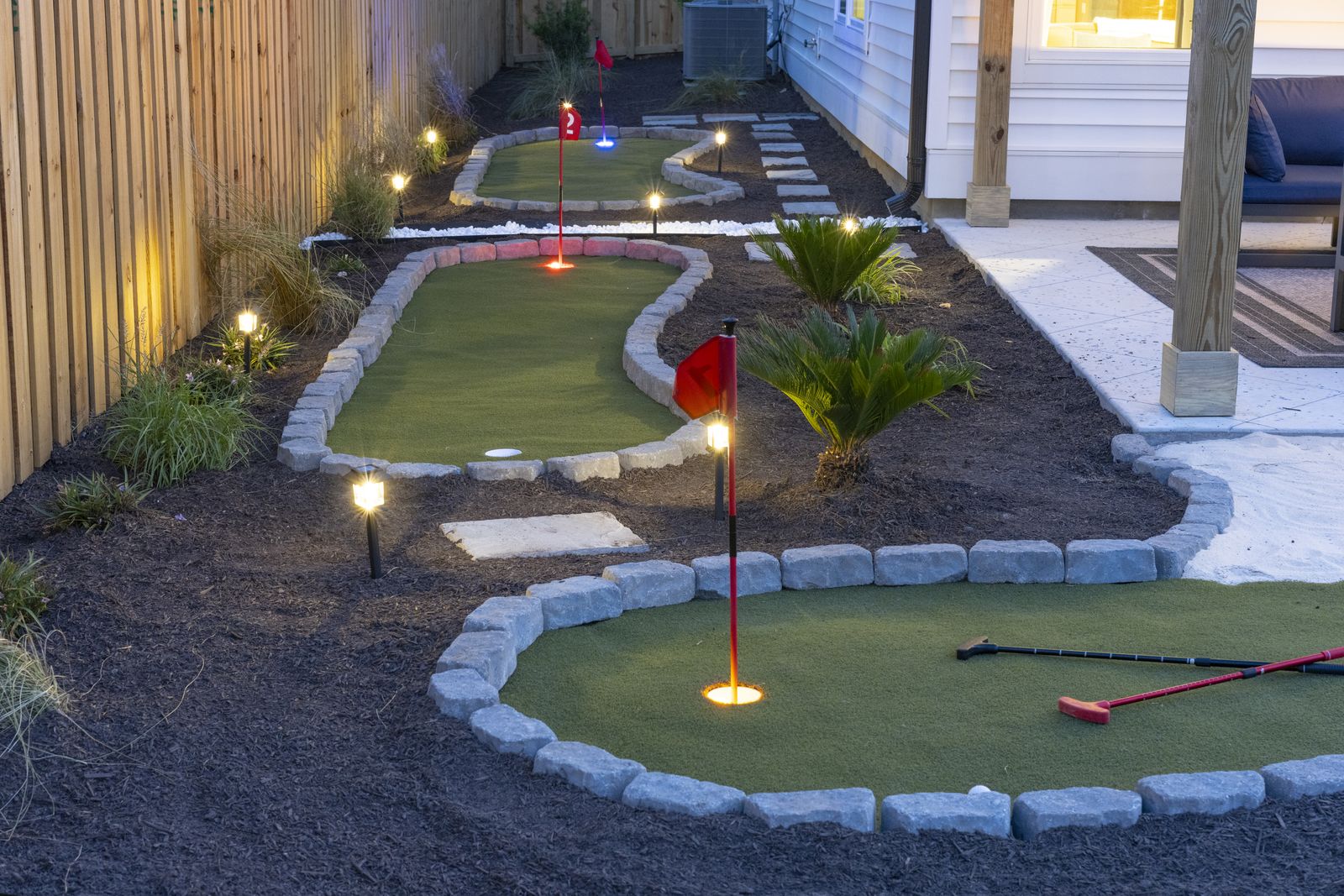 Backyard putting greens lit by path lights at dusk, with flags and putters